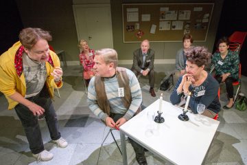 Ryhmä ihmisiä istuu huoneessa puoliympyrän muotoon järjestettynä. Kaksi ihmistä istuu pienen pöydän ääressä, jossa on kynttilöitä ja nimilaput; toinen raitapaitainen mies katsoo takaisin keltaiseen takkiin pukeutuneeseen mieheen, joka puhuu vilkkaasti.