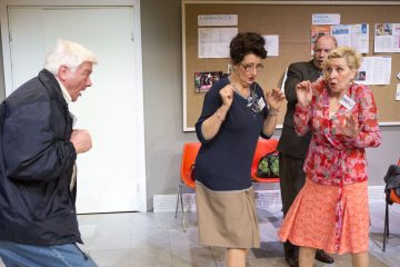 Neljä värikkäisiin vaatteisiin pukeutunutta aikuista seisoo sisätiloissa ilmoitustaulun edessä. He näyttävät käyvän vilkasta tai hätääntynyttä keskustelua, ja heidän ilmeensä ja ruumiinkielensä ovat eloisia.