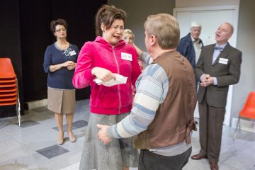 Vaaleanpunaiseen takkiin pukeutunut, silminnähden järkyttynyt nainen seisoo nenäliina kädessään, kun mies kohtaa hänet. Taustalla on neljä ihmistä, joista osa vaikuttaa huolestuneilta. Vasemmalla on pinottu oransseja tuoleja.