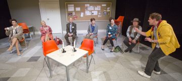 Seitsemän aikuista istuu oransseilla tuoleilla puoliympyrän muotoon, ja yksi mies seisoo ja elehtii vilkkaasti. Taustalla näkyy ilmoitustaulu ja pinottuja tuoleja, jotka viittaavat ryhmäkokoukseen tai harjoitusympäristöön.