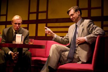 Kaksi pukuihin pukeutunutta miestä istuu punaisilla tuoleilla hämärässä huoneessa; toinen näyttää vakavalta, kun taas toinen osoittaa kädellään kuin keskustelun aikana. Pöydällä on juoma ja tuhkakuppi.