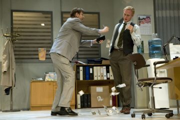 Kaksi liikeasuun pukeutunutta miestä riitelee toimistossa, kun toinen heittelee toista käteisellä. Dollarin setelit putoavat lattialle; taustalla näkyy vedenjäähdytin, tuoli ja toimistotarvikkeita.