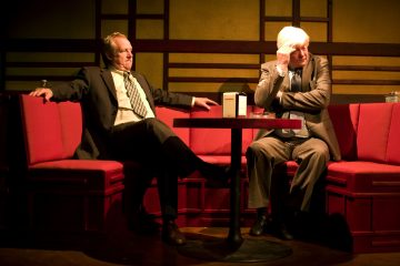 Kaksi pukuihin pukeutunutta miestä istuu punaisilla koppeilla pyöreän pöydän ääressä hämärässä ympäristössä, mahdollisesti baarissa tai ravintolassa. Toinen mies vaikuttaa rentoutuneelta, kun taas toinen näyttää mietteliäältä ja nojaa päätään käteensä.