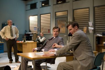 Neljä pukumiestä on toimistossa, jossa on pöytiä, papereita ja tietokoneita. Kaksi miestä istuu ja puhuu pöydän ääressä, yksi seisoo lähellä ja toinen istuu taustalla. Tunnelma vaikuttaa jännittyneeltä ja vakavalta.