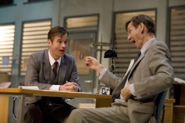 Kaksi pukumiestä istuu vastakkain toimiston pöydän ääressä ja käy vilkasta keskustelua. Toinen mies nojaa eteenpäin hymyillen, kun taas toinen mies elehtii ilmeikkäästi kädellään.