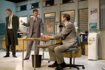 Kolme pukumiestä on toimistolla. Yksi seisoo liitutaulun ääressä, toinen tekee eleitä puhuessaan ja kolmas istuu työpöydän ääressä. Taustalla näkyy toimistokalusteita, papereita ja vedenjäähdytin.