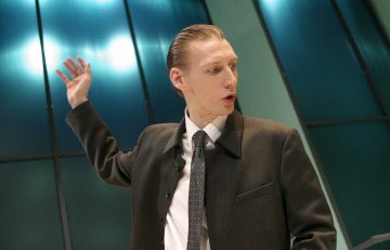 Pukuun ja solmioon pukeutunut mies tekee eleitä nostamalla toisen kätensä ylös ja seisoo suurten, sinivihreiden lasipaneelien edessä, joiden takana heijastuu kirkkaita valoja.