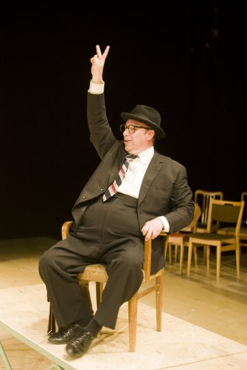 Mustaan pukuun ja hattuun pukeutunut mies istuu puisella tuolilla ja nostaa oikean kätensä kaksi sormea ojennettuna rauhan tai voiton merkiksi. Taustalla lavalla on useita tyhjiä tuoleja.