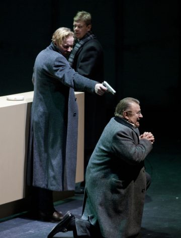 Pitkätakkinen mies osoittaa aseella toista miestä, joka polvistuu ja puristaa kätensä yhteen, kun taas kolmas mies seisoo taustalla, ja kaikilla on tummat takit hämärästi valaistulla näyttämöllä.