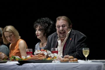 Kolme ihmistä istuu pöydän ääressä, jossa on ruokalautasia ja juomia. Etualalla oleva mies, jolla on silmälasit ja tumma takki, nauraa. Hänen vieressään olevalla naisella on tummat hiukset ja dramaattinen meikki. Asetelma vaikuttaa juhlalliselta.