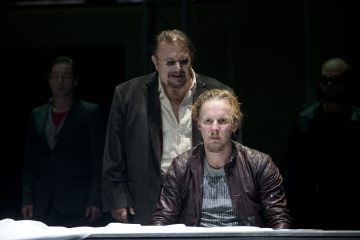 Vakavan näköinen vaaleatukkainen mies, jolla on nahkatakki, istuu pöydän ääressä, ja hänen takanaan seisoo vanhempi mies, jolla on silmälasit ja tumma takki; hämärässä taustalla on kaksi muuta miestä.