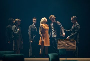 Kuusi ihmistä seisoo hämärästi valaistulla näyttämöllä. Oranssiin mekkoon pukeutunut nainen kohtaa nahkatakkisen miehen, jolle toinen pukumies ojentaa matkalaukkua. Kolme muuta tarkkailee taustalla.