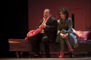 Kaksi näyttelijää istuu näyttämöllä sängyn reunalla dramaattisessa lämpimässä valaistuksessa. Mies pitelee tyynyä ja katsoo mietteliäästi eteenpäin, kun taas takkiin ja kirkkaan vaaleanpunaisiin sukkiin pukeutunut nainen istuu hänen vieressään mietteliäänä.