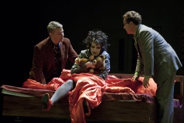 Lavalla on kolme ihmistä. Tummakiharainen nainen istuu punaisella peitolla päällystetyllä sängyllä pehmoleluja kädessään, ja kaksi pukumiestä seisoo hänen kummallakin puolellaan ja vaikuttaa huolestuneilta.