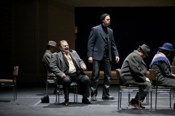 Useita takkeihin ja hattuihin pukeutuneita miehiä istuu näyttämötuoleilla hämärässä teatterikuvassa, kun taas yksi mies seisoo ja katsoo eteenpäin. Tunnelma vaikuttaa vakavalta ja dramaattiselta.