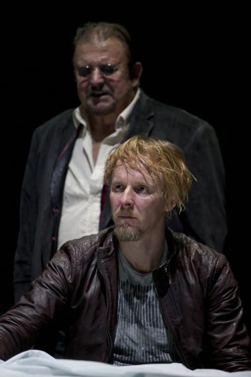 Kuvassa on kaksi miestä mustaa taustaa vasten. Edessä olevalla miehellä on vaaleat hiukset, parta ja ruskea nahkatakki. Takana olevalla vanhemmalla miehellä on silmälasit, lyhyet hiukset ja tumma takki valkoisen paidan päällä.