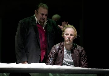 Tummiin takkeihin ja aurinkolaseihin pukeutunut mies seisoo toisen istuvan vaaleatukkaisen miehen takana, joka katsoo juhlallisesti eteenpäin. Seisova mies nostaa kätensä toisen pään lähelle dramaattisessa hämärässä valaistuksessa.