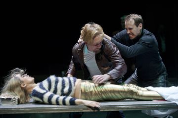 Kolme näyttelijää esittää dramaattisen kohtauksen: raidallinen pusero ja kultaiset housut yllään oleva nainen makaa pöydällä, kun kaksi miestä kamppailee hänen vieressään, toinen tarttuu toisen päähän, ja molemmat vaikuttavat intensiivisiltä ja tunteikkailta.