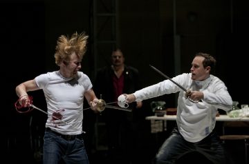 Kaksi miestä käy lavalla kiivasta miekkataistelua, ja toisen valkoinen paita on veressä. Kolmas henkilö seisoo taustalla ja katsoo kaksintaistelua. Kohtaus vaikuttaa dramaattiselta ja teatraaliselta.