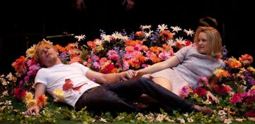 Mies, jolla on verinen valkoinen paita, ja nainen harmaassa mekossaan makaavat kädestä pitäen värikkäiden tekokukkien keskellä, vehreiden kasvien ympäröimänä, ja molemmat katsovat sivulle.