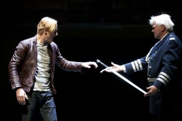 Kaksi miestä lavalla; toinen nahkatakkinen mies kurottautuu kohti miekkaa, jota pitelee toinen mies, jolla on tumma, hopealla koristeltu univormu. Tausta on tumma, mikä kiinnittää huomion heidän vuorovaikutukseensa.