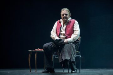 Lavalla istuu tuolilla mies, jolla on valkoinen paita ja punainen liivi, kädessään tumma takki. Hänen vieressään on pieni pöytä, jossa on papereita tai esineitä. Tausta on tumma, mikä korostaa dramaattista kohtausta.