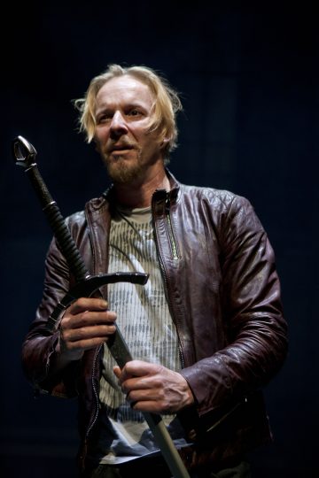 Vaaleatukkainen mies, jolla on ruskea nahkatakki ja raidallinen paita, pitää suurta miekkaa pystyssä molemmissa käsissään seisoen tummaa taustaa vasten.