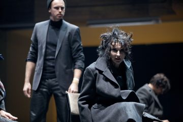 Lavalla esiintyy kaksi näyttelijää; toinen istuu tummassa takissa, hiukset sotkuiset ja ilme on ahdistunut, kun taas toinen seisoo takana puku päässään ja pipo päässään, molemmat dramaattisessa valaistuksessa.