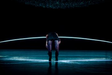 Henkilö seisoo pimeällä näyttämöllä eteenpäin kumartuneena, kädet roikkuvat alaspäin, ja taustalla näkyy kaareva sinisen valon viiva ja heijastuksia lattialla, mikä luo dramaattisen, minimalistisen ilmapiirin.