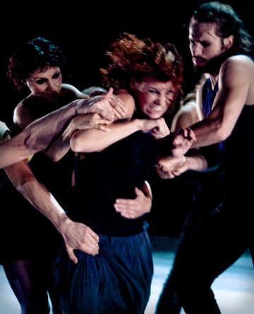 Kolme ihmistä tarttuu eri kulmista naiseen, joka kamppailee, ja hänen kasvonsa ovat jännittyneet tunteista. Kohtaus on dramaattinen, ja hämärä valaistus korostaa heidän fyysisen vuorovaikutuksensa voimakkuutta.