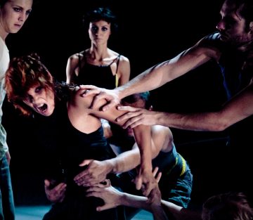 Viisi ihmistä on pimeällä näyttämöllä. Yksi edessä oleva henkilö vaikuttaa ahdistuneelta, kun muut kurottautuvat ja tarttuvat hänen käsiinsä ja hartioihinsa. Taustalla oleva henkilö katselee vakavalla ilmeellä. Kohtaus on jännittynyt ja dramaattinen.