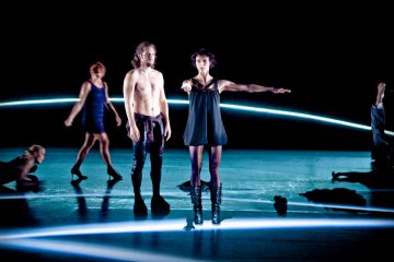 Ryhmä tanssijoita esiintyy lavalla, jota valaisevat sinivalkoiset neonvalot. Paidaton mies ja mustapukuinen nainen seisovat keskellä; muut poseeraavat tai liikkuvat taustalla.