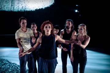 Kuusi esiintyjää seisoo hämärästi valaistulla näyttämöllä, edessä yksi nainen, kädet ojennettuina ja ilmeikäs ilme kasvoillaan. Takana oleva ryhmä katsoo tarkkaavaisesti. Yläpuolella roikkuu abstrakti, verkkomainen rakenne.