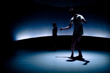 Kaksi ihmistä seisoo hämärästi valaistulla näyttämöllä; toinen etualalla poseeraa kädet ojennettuina ja heittää varjon, kun taas toinen seisoo kauempana takana, ja molempia ympäröi dramaattinen sininen valaistus.