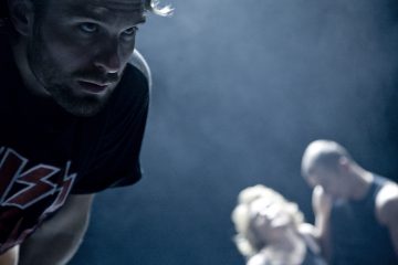 Parrakas mies on tarkennettu ja katsoo intensiivisesti vasemmalle; kaksi epäselvää hahmoa, toinen vaalea ja toinen kalju, on sumuisessa taustassa. Valaistus on hämärä ja sinisen sävyinen.