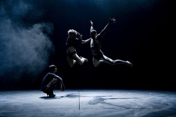 Lavalla esiintyy kolme tanssijaa, joista kaksi hyppää ilmassa ojennetuin käsin ja jaloin, kun taas yksi kyykistelee lattialla. Dramaattinen valaistus ja sumu luovat tunnelmallisen, teatraalisen ilmapiirin.