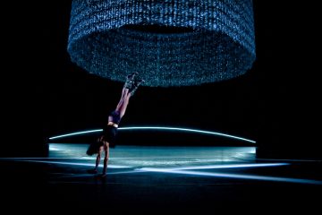 Esiintyjä tekee käsilläseisontaa lavalla suuren, valaistun, pyöreän, sinisen installaation alla, jossa on ryöppyäviä säikeitä. Kohtaus on dramaattisesti valaistu sinisellä valolla ja hehkuvilla viivoilla lattialla.