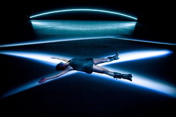 Mustaan mekkoon ja saappaisiin pukeutunut henkilö makaa selällään kädet ja jalat ojennettuina keskellä dramaattisia sinisiä ja valkoisia valonsäteitä, jotka risteävät pimeällä näyttämöllä. Taustalla loistaa kaareva sininen valo.