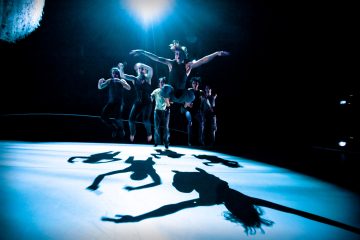 Tanssijat hyppäävät lavalla dramaattisessa sinisessä valaistuksessa, joka heittää suuria, ilmeikkäitä varjoja heidän taakseen, ja yhdellä tanssijalla on etualalla kukkainen päähine.