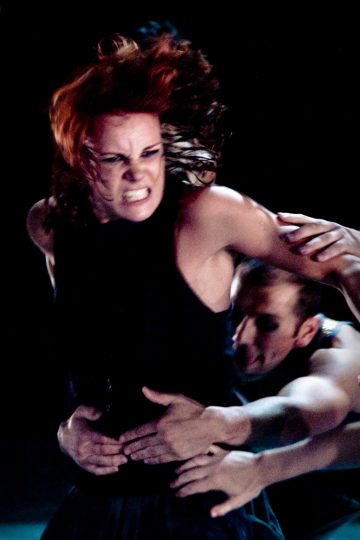 Punatukkainen, hurja ilmeinen nainen, jota mies pitää tiukasti vyötärön ympärillä, molemmat tummiin vaatteisiin pukeutuneina, tummalla taustalla, mikä luo dramaattisen ja intensiivisen kohtauksen.