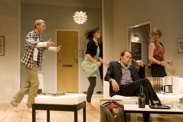 Neljä ihmistä on modernissa olohuoneessa. Kolme liikkuu tai tanssii tarmokkaasti, kun taas yksi pukumies istuu sohvalla ja näyttää väsyneeltä tai välinpitämättömältä. Pöydällä on pullo, lasit ja laukku.