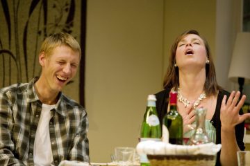 Mies ja nainen istuvat pöydässä juomien ääressä nauraen ja hymyillen. Mies nauraa silmät kiinni, kun taas nainen nojaa taaksepäin, silmät kiinni ja käsi rinnalla, ilmaisten huvittuneisuutta tai iloa.