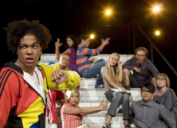 Joukko nuoria aikuisia poseeraa teatterimaisesti valkoisilla portailla, jotkut istuvat ja jotkut elehtivät dramaattisesti. Etualalla on värikkääseen takkiin pukeutunut henkilö, joka tekee ilmeikkäitä ilmeitä. Taustalla loistavat näyttämön valot.