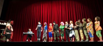 Ryhmä värikkäisiin, eklektisiin asuihin pukeutuneita ihmisiä seisoo lavalla punaisen verhon edessä. Vasemmalla nainen istuu tuolissa ja lapsi näppäimistön ääressä. Muu näyttelijäkaarti on heitä vastapäätä näyttämön toisella puolella.