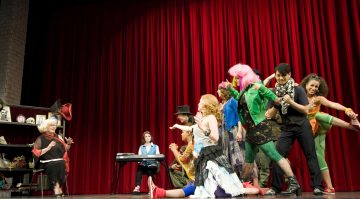 Ryhmä värikkäästi pukeutuneita esiintyjiä toimii energisesti lavalla punaisen verhon edessä, kun nainen istuu näppäimistön ääressä ja toinen nainen tekee dramaattisia eleitä tuolilta rekvisiittaa sisältävän hyllyn vieressä.