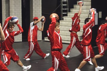 Joukko punavalkoisiin urheiluasuihin pukeutuneita ihmisiä suorittaa sisätiloissa koreografioidun harjoituksen, jossa jokaisella on koripallo kädessään ja nostaa sen päänsä yläpuolelle. Taustalla näkyvät valkoiset kaakeliseinät ja portaat.