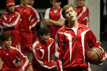 Punaisiin verkkareihin pukeutunut nuori mies pitää koripalloa kädessään ja näyttää laulavan tai puhuvan intohimoisesti, kun taustalla katselee useita punaisiin verkkareihin pukeutuneita joukkuetovereita.