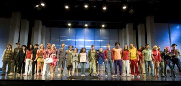 Monipuolinen joukko esiintyjiä seisoo vierekkäin lavalla värikkäisiin, eklektisiin asuihin pukeutuneena kirkkaiden valojen alla ja näyttää kumartavan teatteriesityksen päätteeksi.