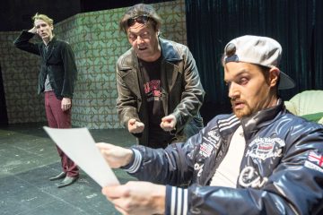 Kolme miestä näyttelee näyttämöllä intensiivistä kohtausta; yksi istuu lippalakkiin ja takkiin pukeutuneena ja yllättyneenä kädessään pitelemästään paperista, kun taas kaksi muuta seisovat takana, toinen turhautuneena ja toinen hämmentyneenä.