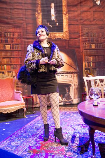 Leopardikuvioiseen takkiin, mustaan mekkoon, verkkosukkiin ja korkeakorkoisiin saappaisiin pukeutunut nainen seisoo itsevarmasti lavasteessa, joka muistuttaa eleganttia kirjastoa, jossa on vintage-huonekaluja ja takkatausta.